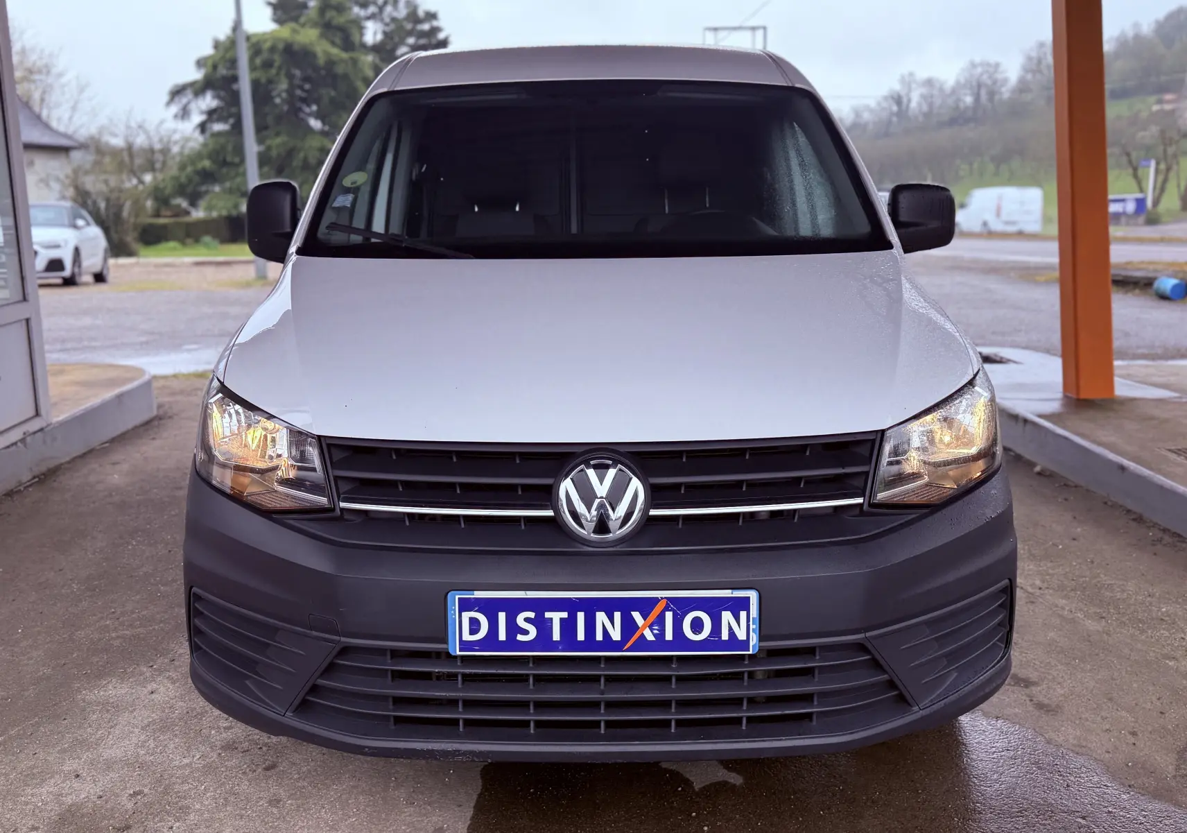 Vue de face d'un Volkswagen Caddy blanc avec pare-chocs noir et phares allumés sous abri.