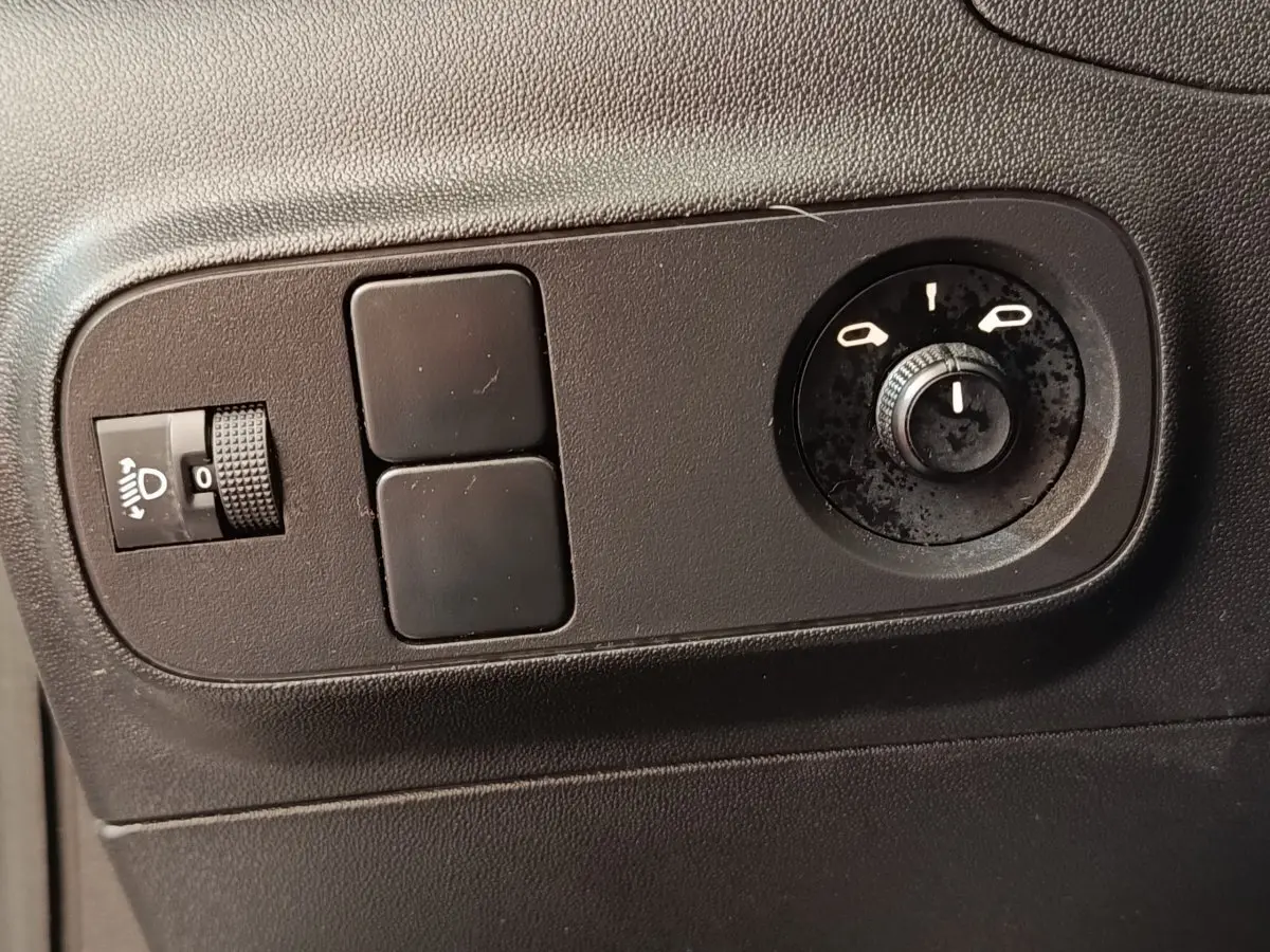 Gros plan sur les commandes noires de réglage des phares et des rétroviseurs d'une Citroën C3 blanche, côté gauche.