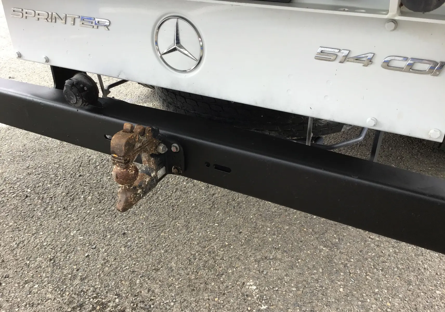 Gros plan sur l'attelage rouillé et le pare-chocs noir du Mercedes Sprinter blanc 514 CDI à l'arrière.