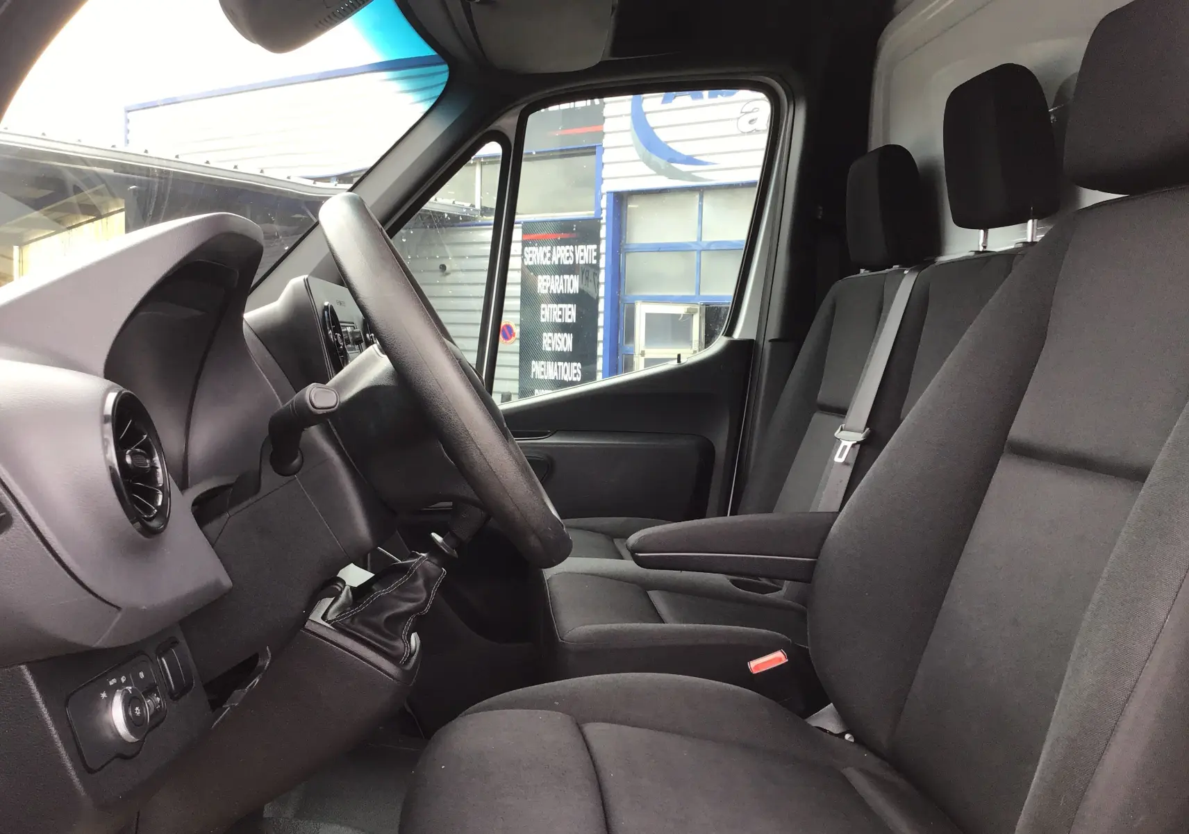 Vue intérieure côté conducteur du Mercedes Sprinter blanc 2018, montrant le volant, levier de vitesses manuel et sièges en tissu gris.