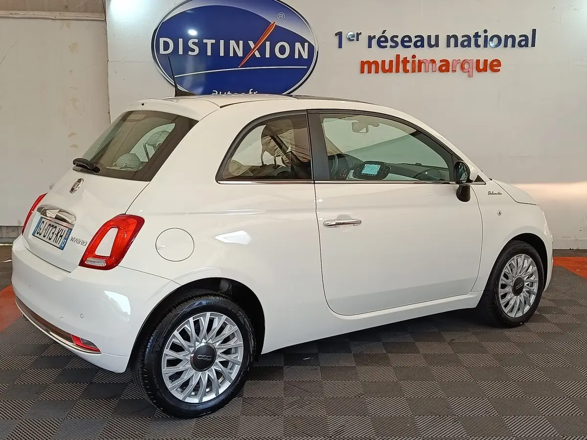 Fiat 500 Hybrid blanc vue 3/4 arrière droit en intérieur, avec jantes alliage et logo Dolcevita sur aile avant.