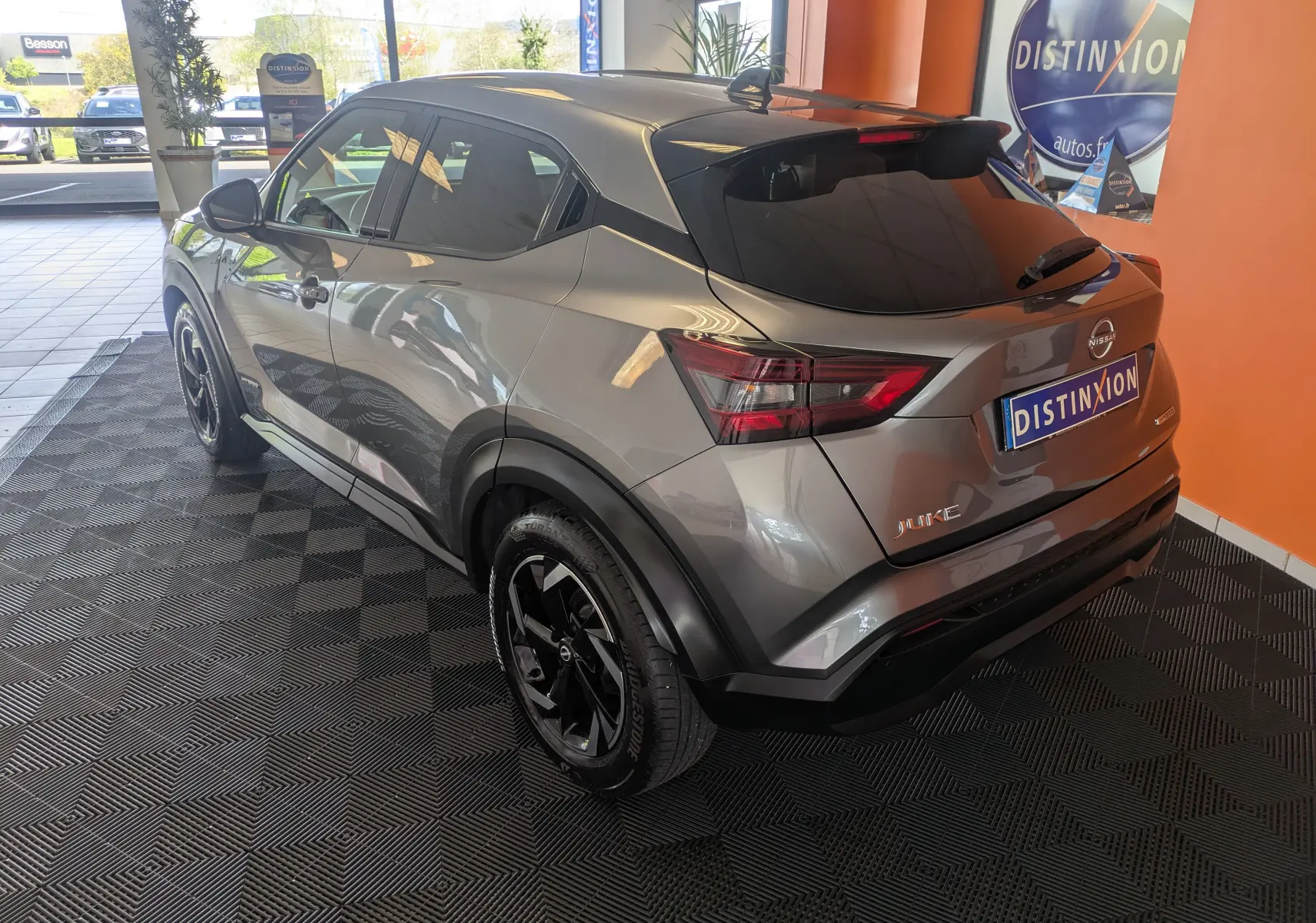 Nissan Juke 2024 gris squale métal en vue 3/4 arrière droit, avec jantes noires et vitres teintées dans un showroom.