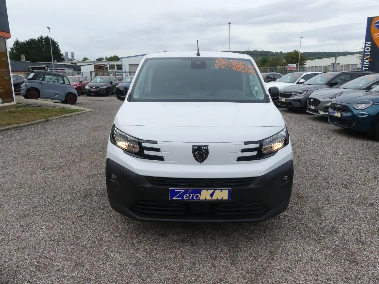 Vue de face d'un Peugeot Partner blanc avec calandre noire et logo lion, stationné sur un parking extérieur.
