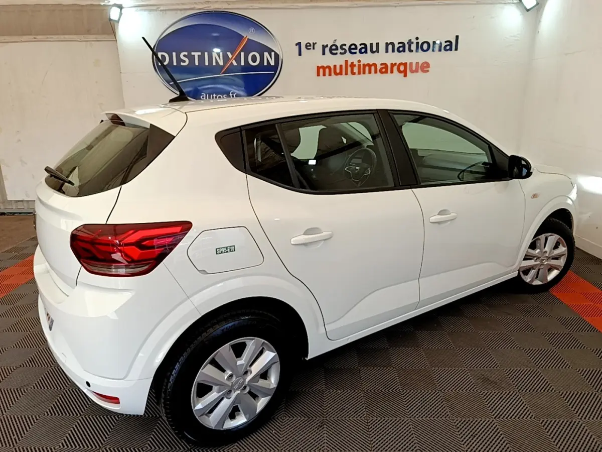 Vue 3/4 arrière droite d'une Dacia Sandero Confort TCe 90 blanche, avec jantes alliage et logo SP95-E10 sur la trappe à carburant.