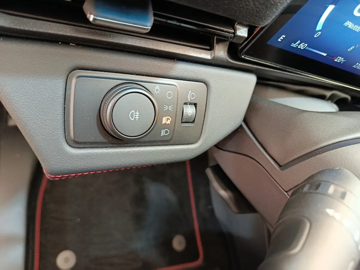 Gros plan sur le bouton de réglage des phares avec éclairage allumé, tableau de bord noir et surpiqûres rouges Ford Puma 2025.