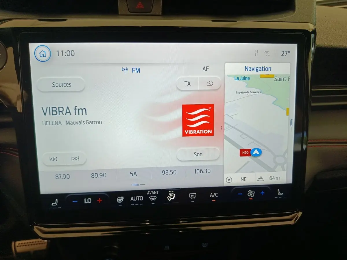 Écran tactile central du Ford Puma 2025 affichant radio et navigation, avec tableau de bord noir et surpiqûres rouges visibles.