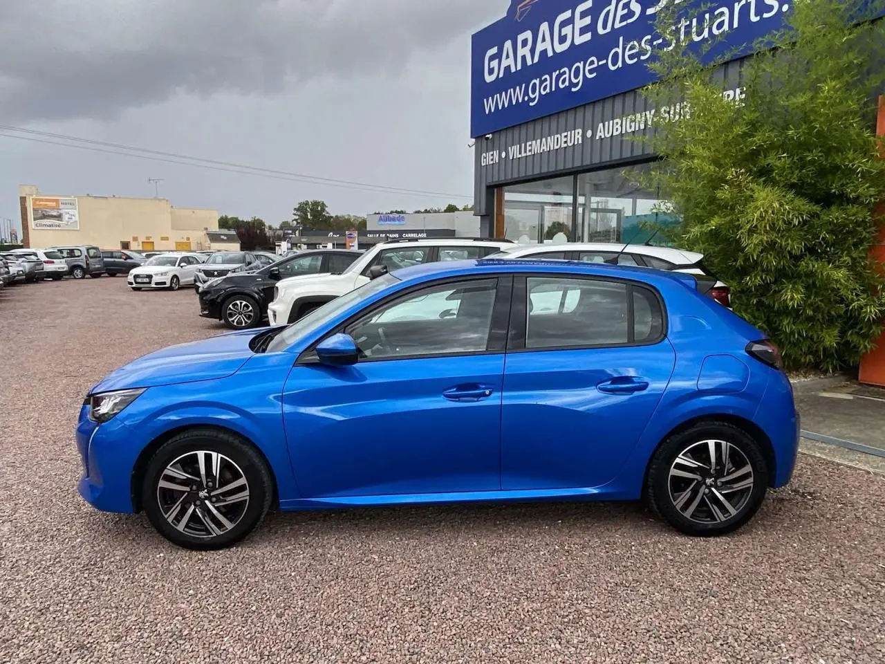 Profil côté gauche d'une Peugeot 208 bleue 2020 avec jantes alliage 16'' et vitres arrière teintées, stationnée devant un garage.
