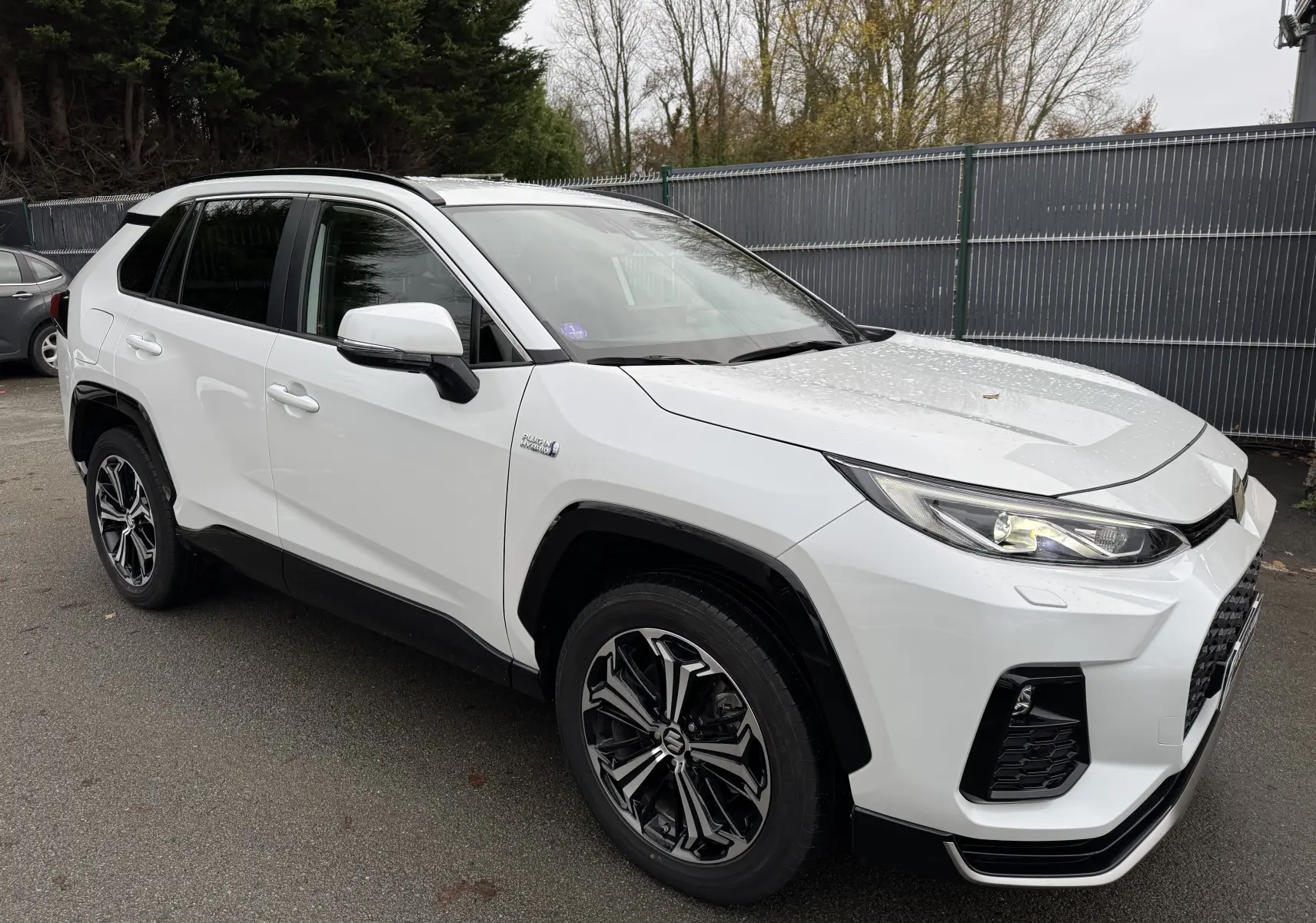 Suzuki Across blanc en vue 3/4 avant droit, avec phares allumés et jantes alliage noires distinctives.