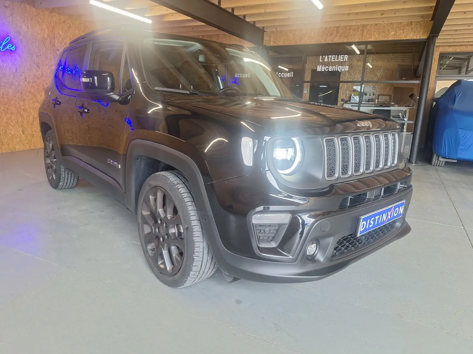 Jeep Renegade noir en 3/4 avant droit avec phares allumés dans un garage moderne.