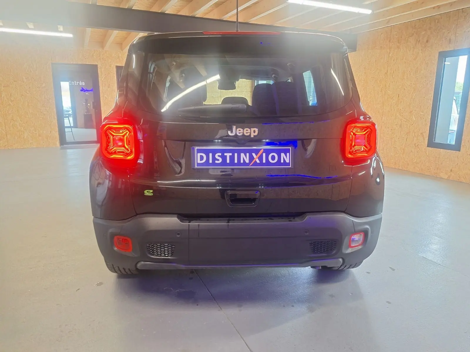 Vue arrière d'un Jeep Renegade noir 2024 avec feux arrière allumés et plaque Distinxion dans un intérieur lumineux.