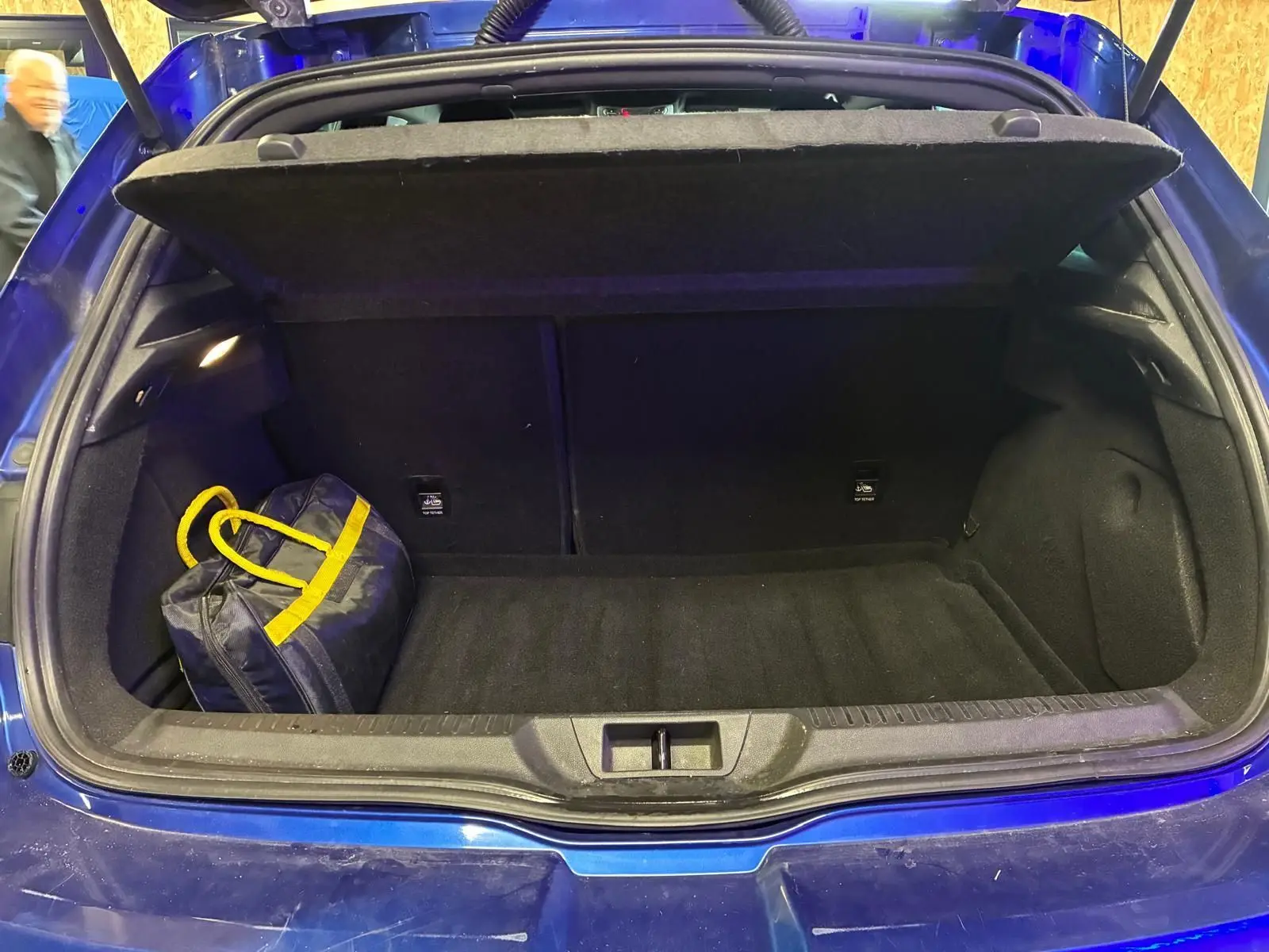 Coffre ouvert d’une Renault Mégane bleu 2019, vue arrière, avec un sac noir à anses jaunes à l’intérieur.