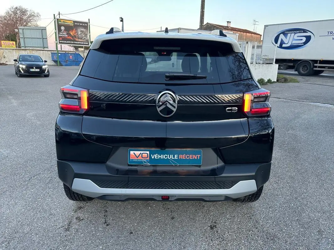 Vue arrière d'une Citroën C3 noire 2025 avec barres de toit et feux arrière allumés sur parking.