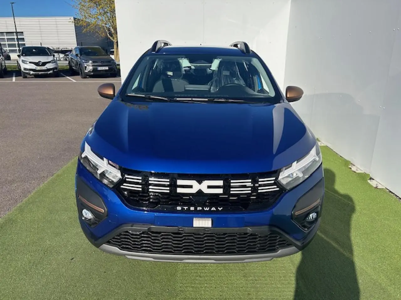 Vue avant d'une Dacia Sandero III Stepway bleu Iron avec calandre noire et détails brun cuivré sur un sol vert.