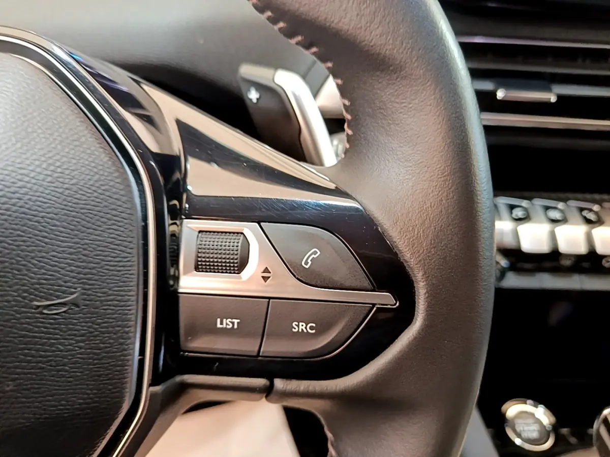 Gros plan sur les commandes gauche du volant cuir noir du Peugeot 3008 gris foncé, avec tableau de bord flou en arrière-plan.