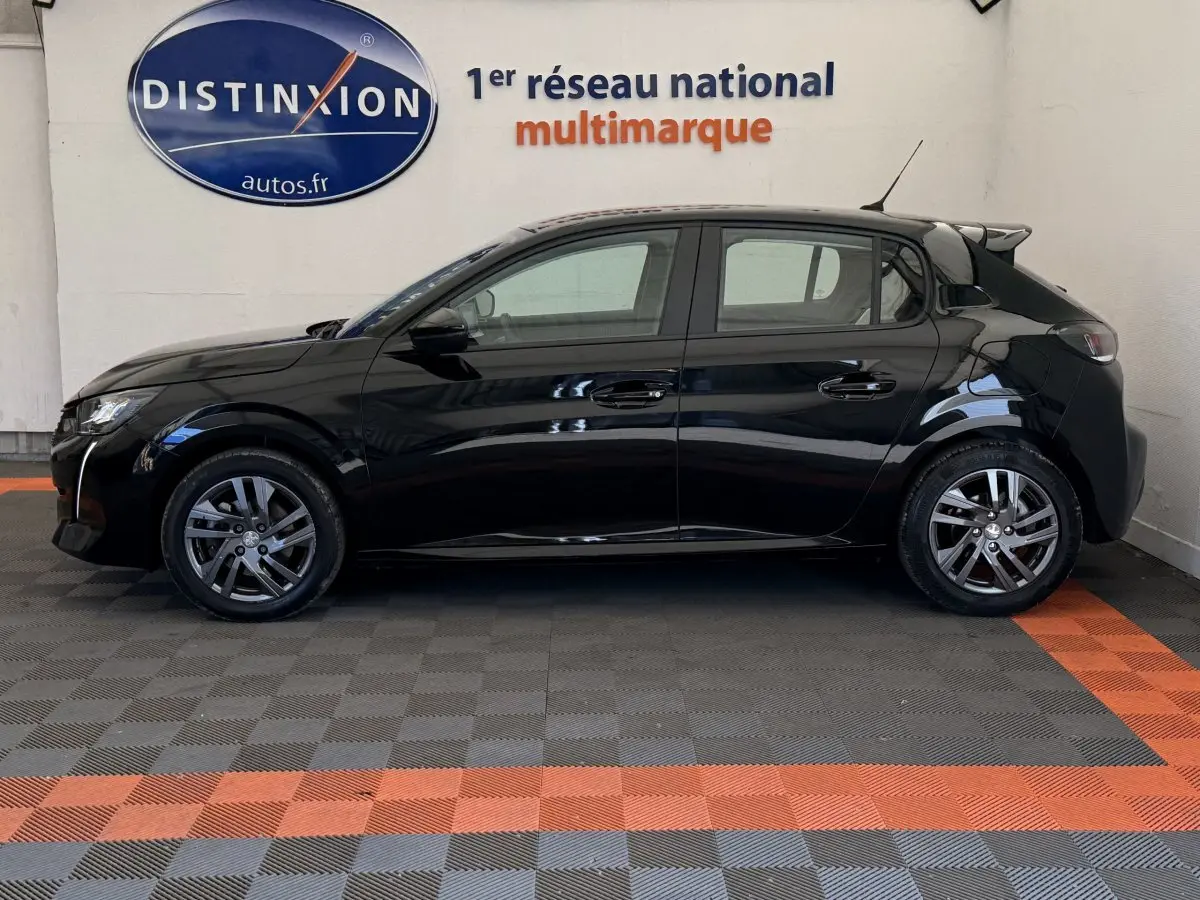 Profil côté gauche d'une Peugeot 208 noire 2021 garée en intérieur sur sol damier gris et orange.