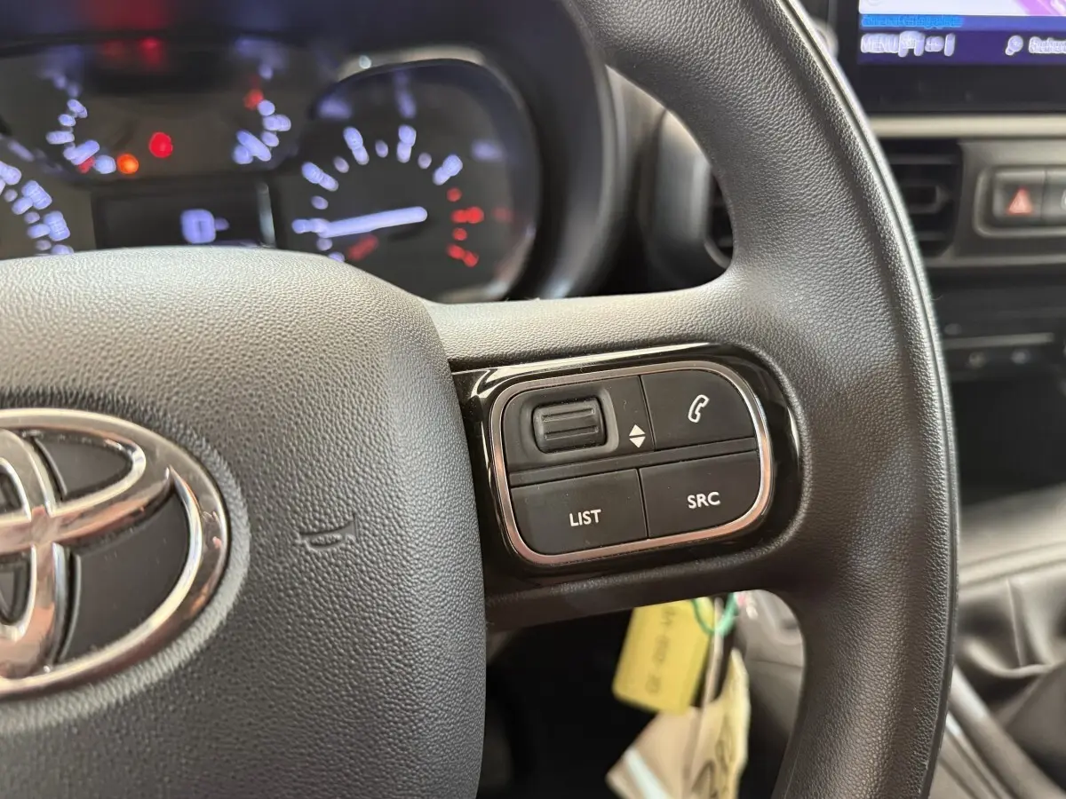 Gros plan sur le volant noir multifonctions de la Toyota Proace City 2022 avec commandes audio et tableau de bord éclairé.