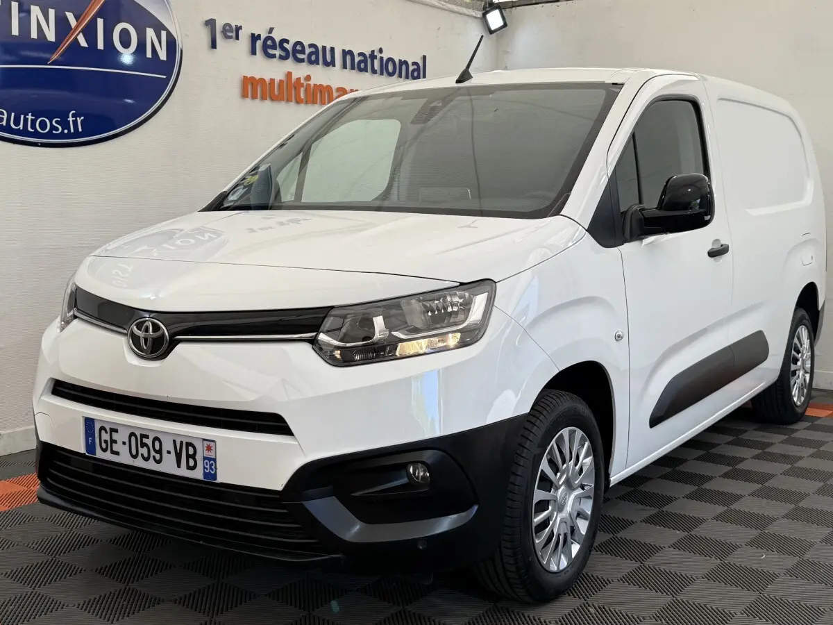 Toyota Proace City blanc en vue 3/4 avant droit, avec calandre noire et phares allumés dans un showroom.