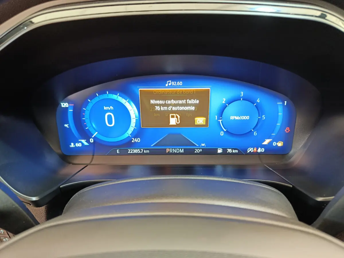 Tableau de bord numérique bleu du Ford Kuga 2024 affichant une alerte de faible carburant, vu de face depuis le volant.