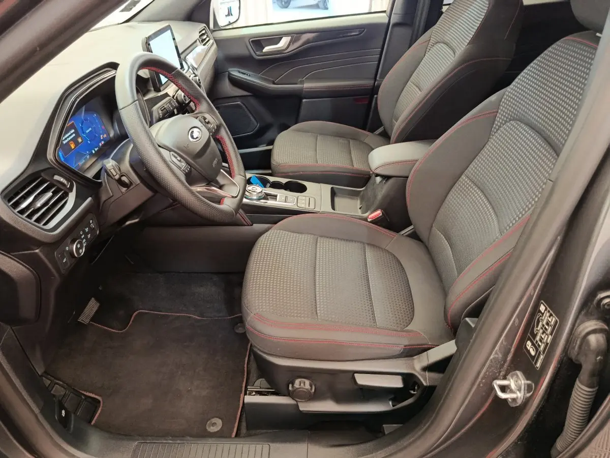 Vue intérieure côté conducteur du Ford Kuga 2024 St Line, sièges tissu gris avec surpiqûres rouges et tableau de bord digital.