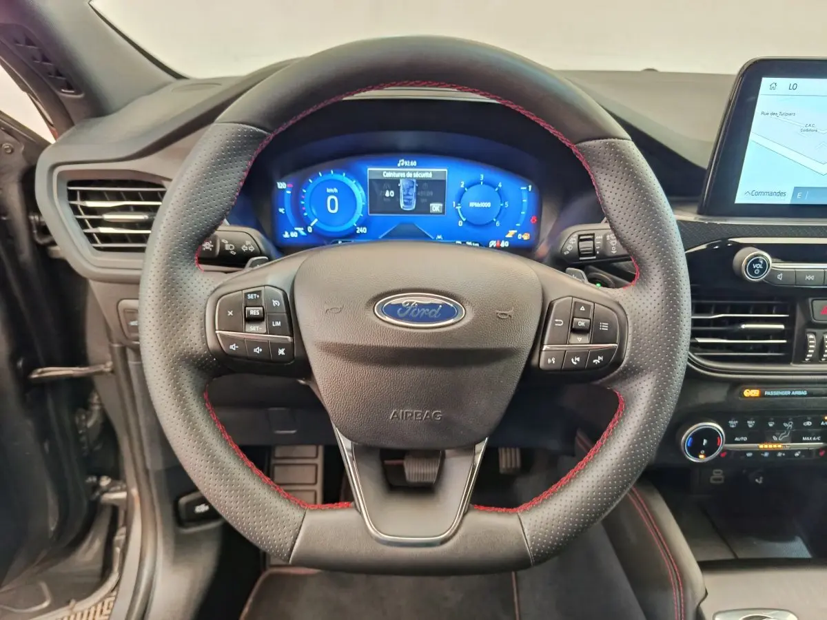 Vue rapprochée du volant cuir noir avec surpiqûres rouges et tableau de bord numérique bleu du Ford Kuga 2024 gris clair.