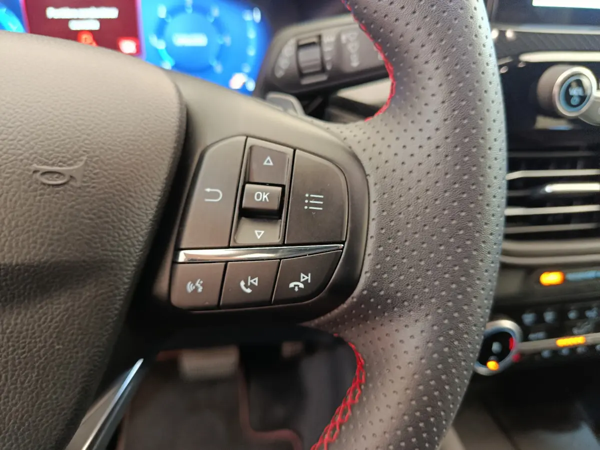 Gros plan sur le volant en cuir perforé avec surpiqûres rouges du Ford Kuga 2024, commandes multifonctions visibles.
