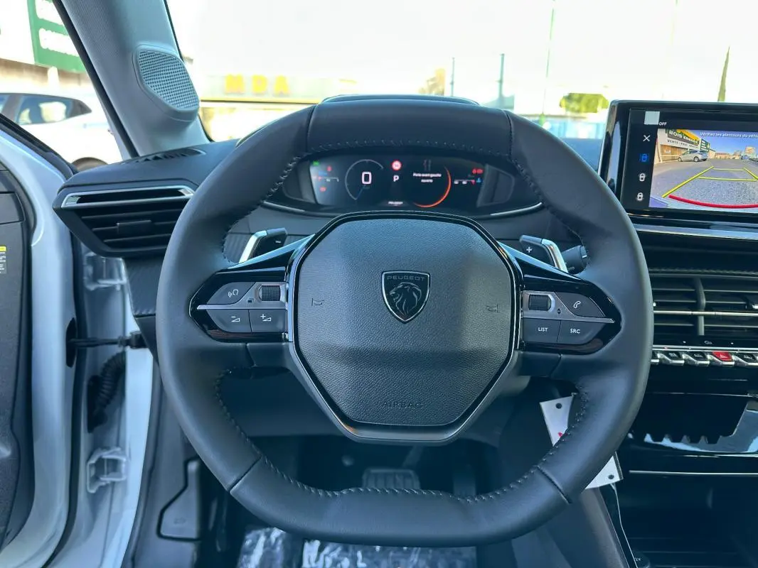 Vue centrée sur le volant noir de la Peugeot 208 blanc 2025, avec tableau de bord numérique et écran tactile arrière en arrière-plan.