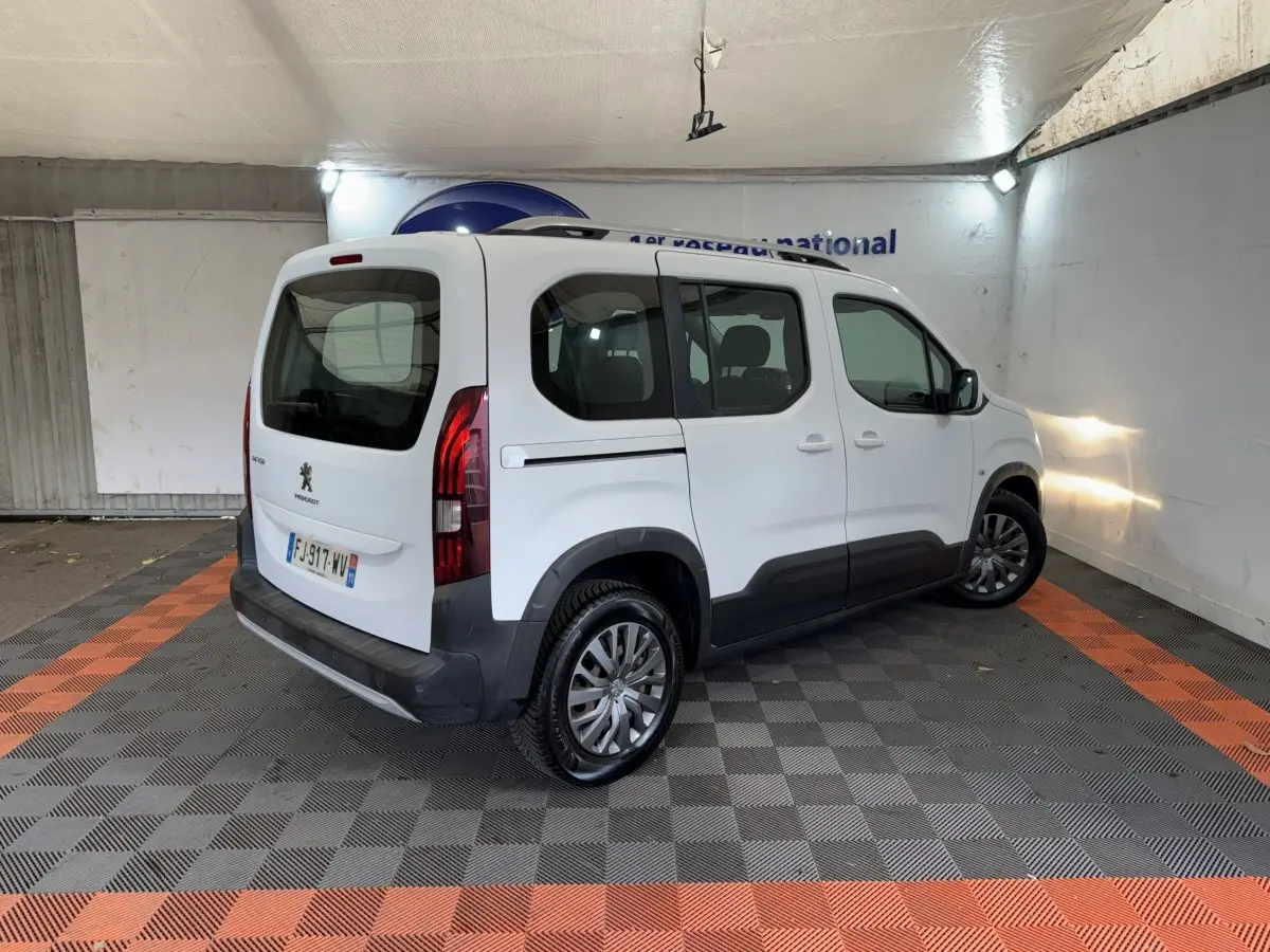 Vue 3/4 arrière droite du Peugeot Rifter blanc avec barres de toit et protections latérales noires dans un garage.