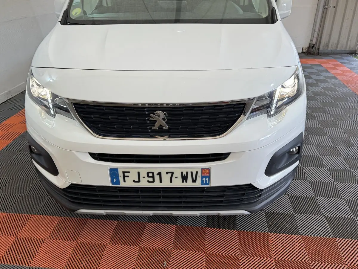Vue avant rapprochée d’un Peugeot Rifter blanc avec calandre noire et phares allumés dans un garage.