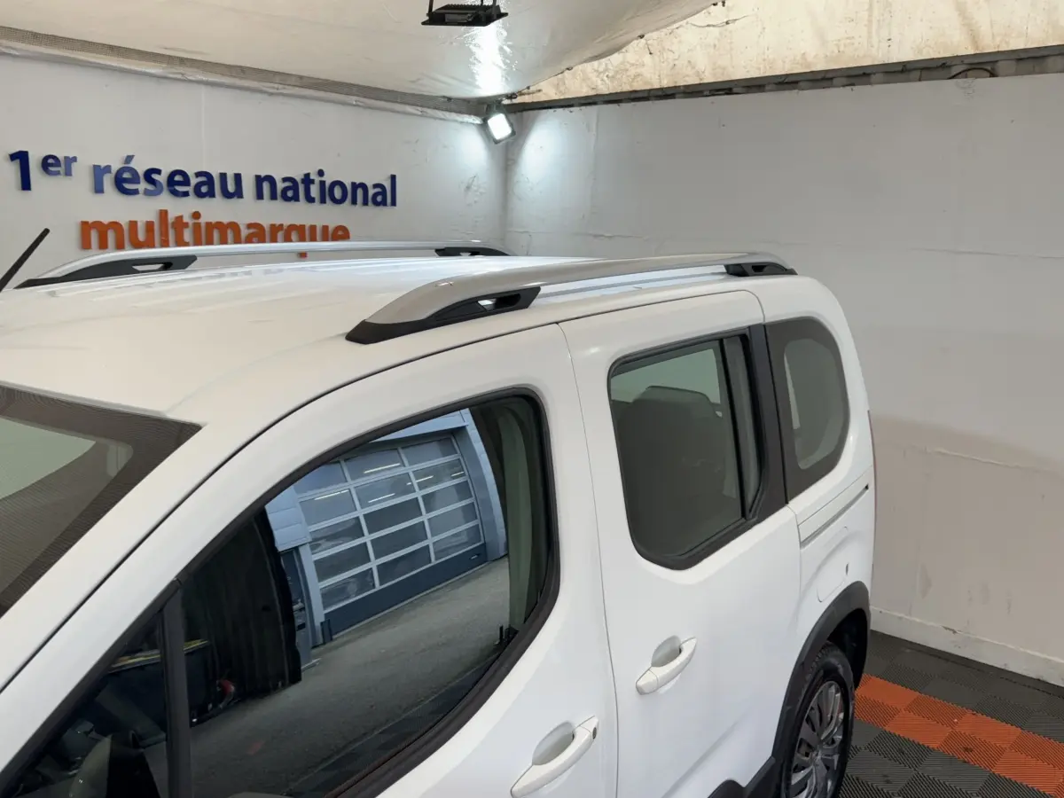 Vue 3/4 arrière droite du Peugeot Rifter blanc avec barres de toit noires et vitres teintées dans un garage.