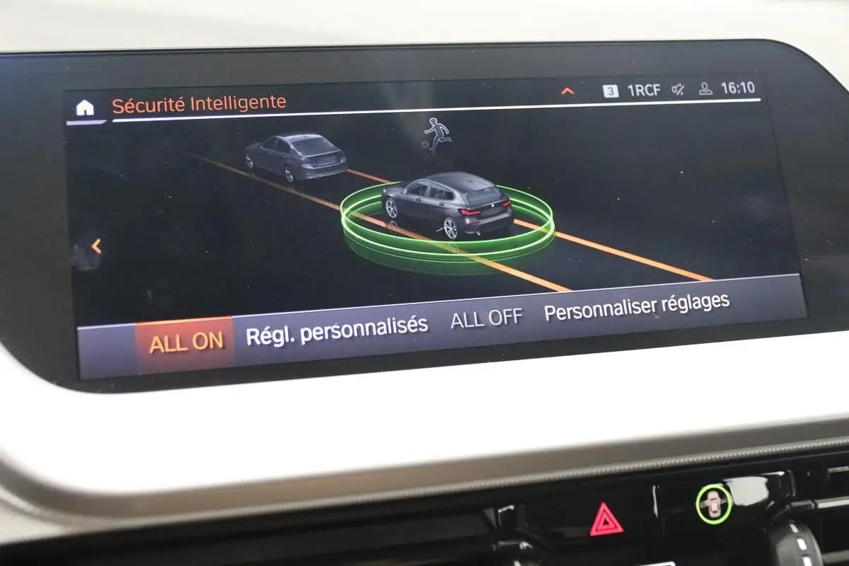 Écran central de la BMW Série 1 16d 2023 montrant l'interface de sécurité intelligente avec vue virtuelle du véhicule noir en circulation.