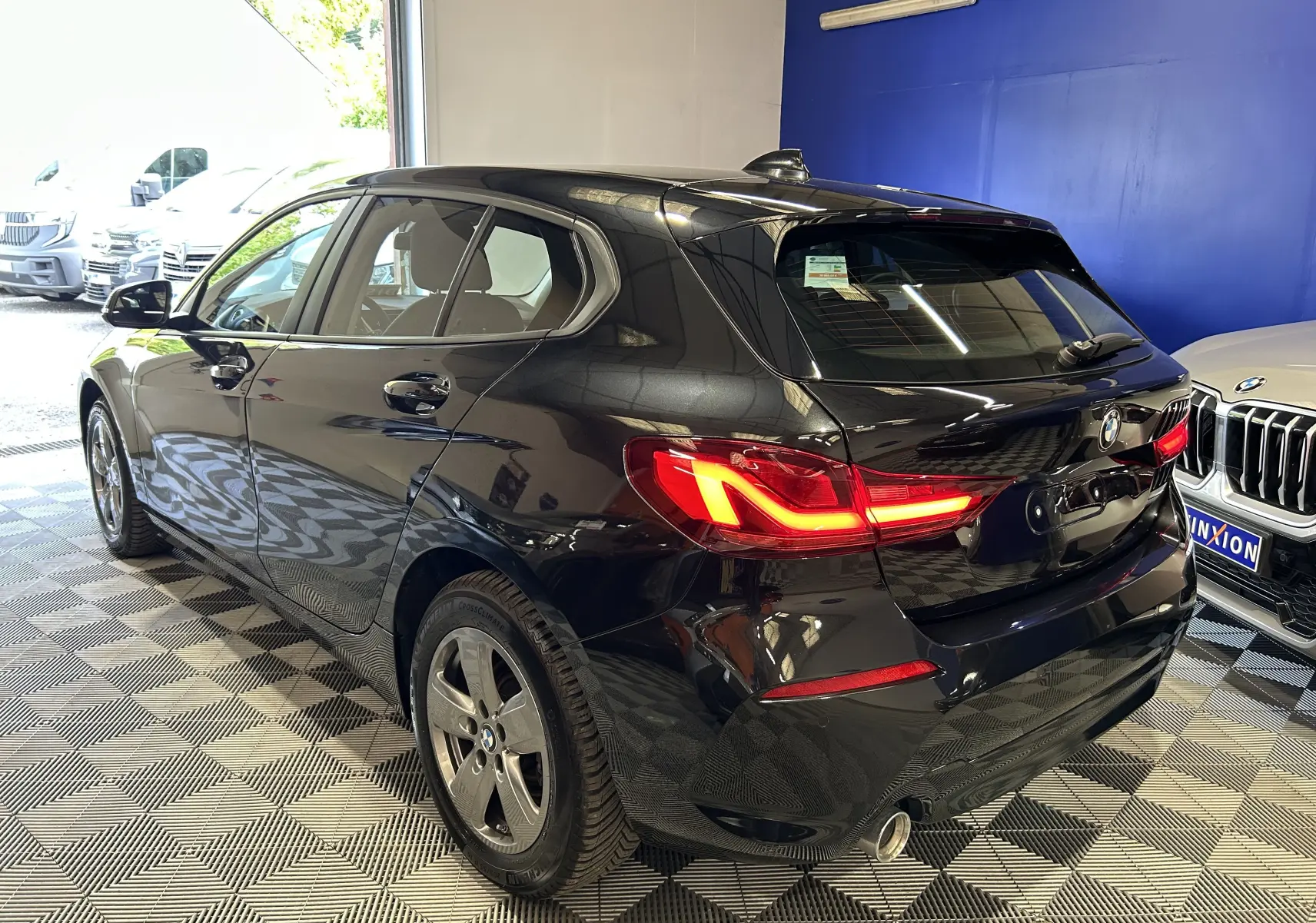 BMW Série 1 16d noire vue en 3/4 arrière droit, feux arrière allumés dans un showroom lumineux.