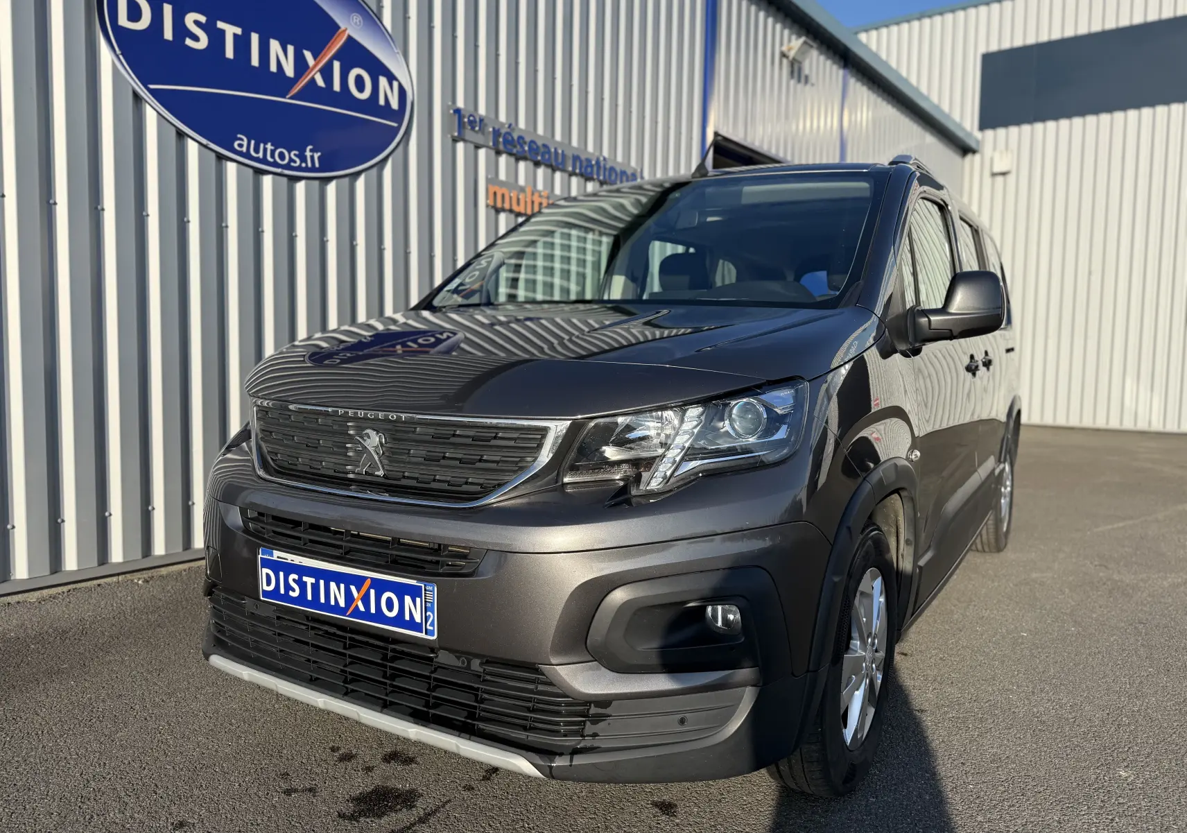 Peugeot Rifter gris platinium en 3/4 avant droit, avec calandre noire et jantes alliage visibles, stationné devant un bâtiment.