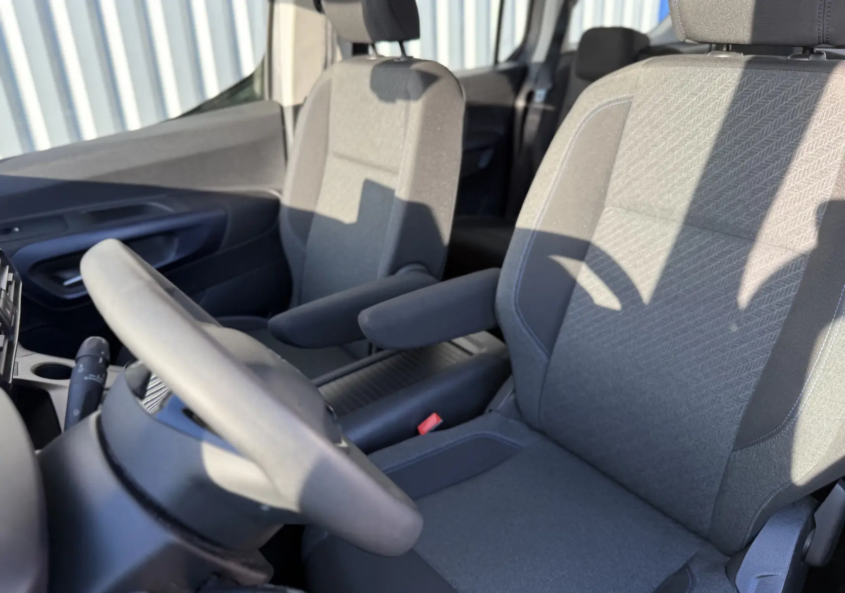 Vue intérieure rapprochée des sièges avant en tissu gris du Peugeot Rifter 1.5 BlueHDi 130, avec accoudoirs et volant partiel.