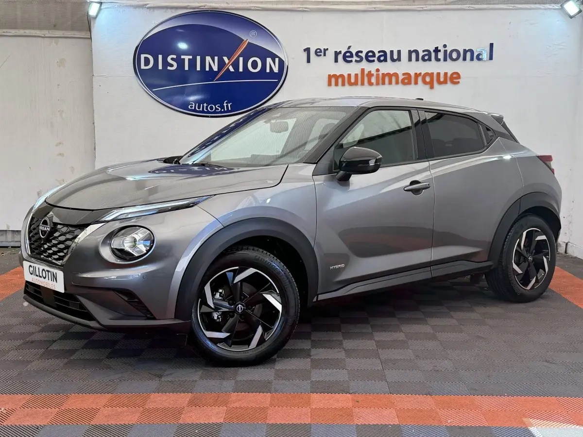 Nissan Juke gris foncé en 3/4 avant droit, avec jantes noires et logo Hybrid visible sur l'aile avant.