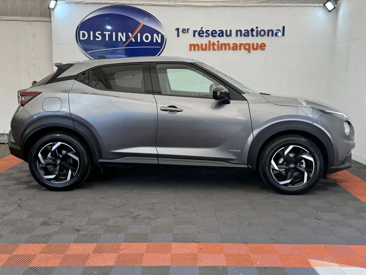 Vue de profil côté gauche d'un Nissan Juke gris foncé 2023 hybride avec jantes noires et logo Distinxion en arrière-plan.