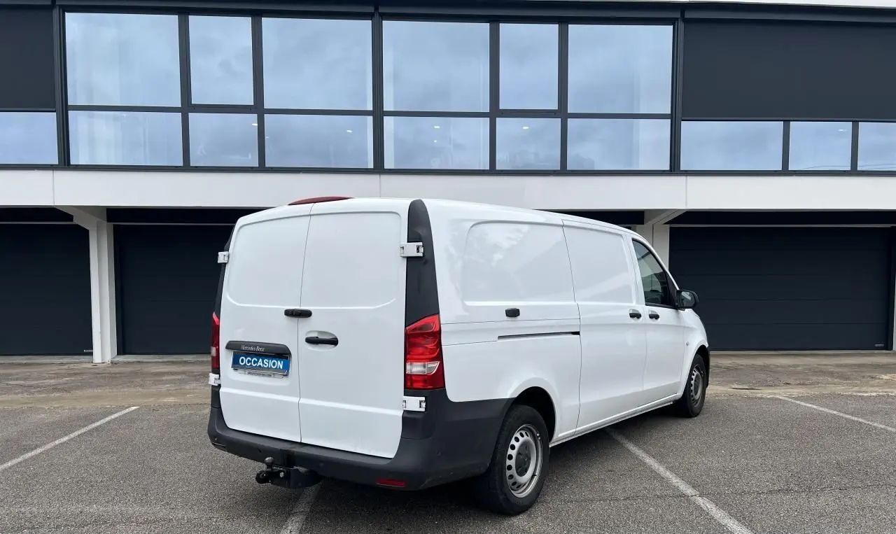 Vue 3/4 arrière droite d’un Mercedes Vito Fourgon blanc avec attelage et porte latérale, stationné devant un bâtiment moderne.