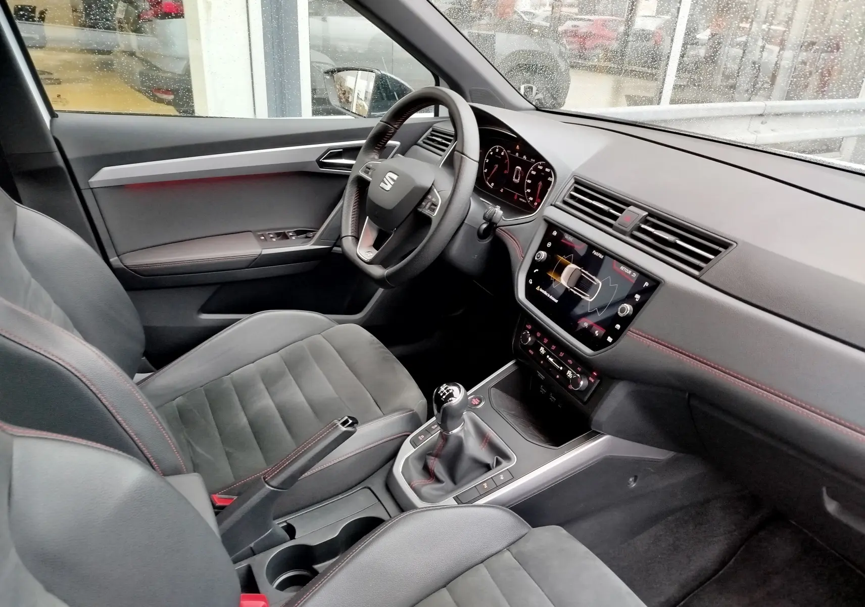 Vue intérieure côté conducteur du SEAT Arona 2020, montrant le volant, la console centrale avec boîte manuelle et les sièges en cuir noir avec surpiqûres rouges.