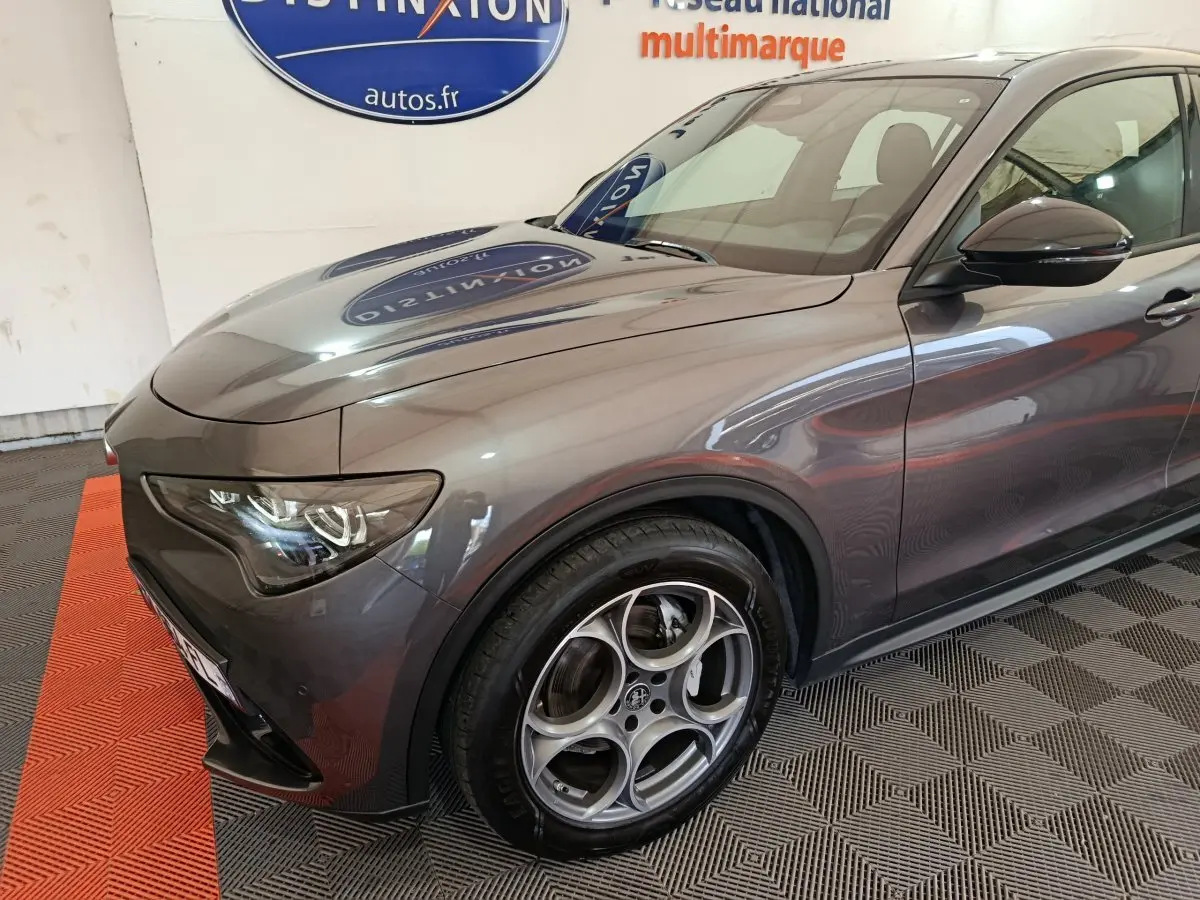 Vue 3/4 avant gauche d'un Alfa Romeo Stelvio gris foncé avec jantes alliage distinctives et phares LED allumés.