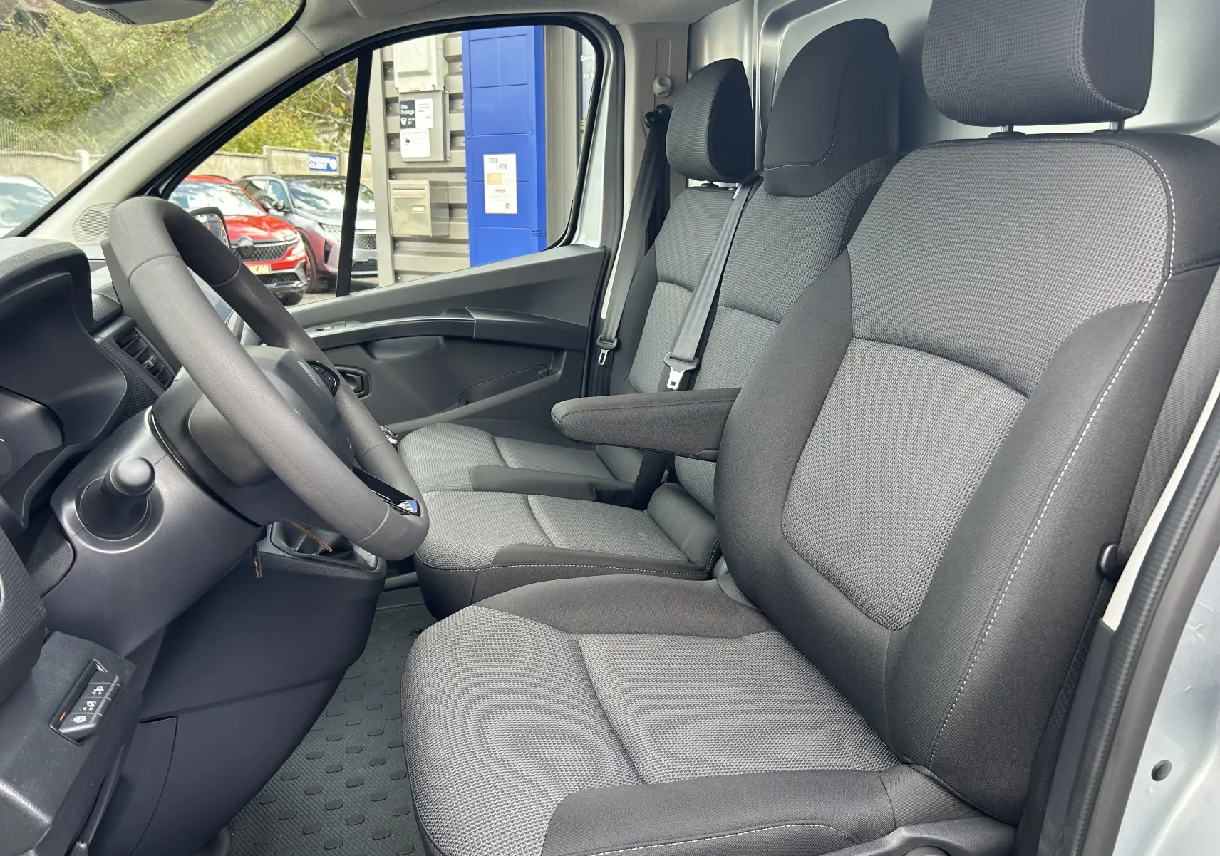 Vue intérieure côté conducteur du Renault Trafic Fourgon blanc, montrant les sièges tissu gris et le volant.