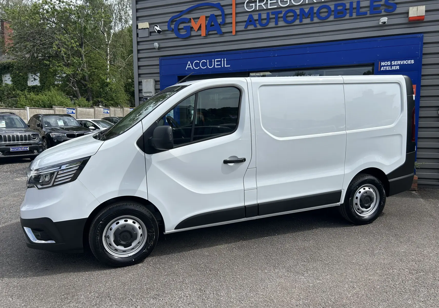 Renault Trafic Fourgon blanc en profil droit, stationné devant un bâtiment commercial avec logo GMA visible.