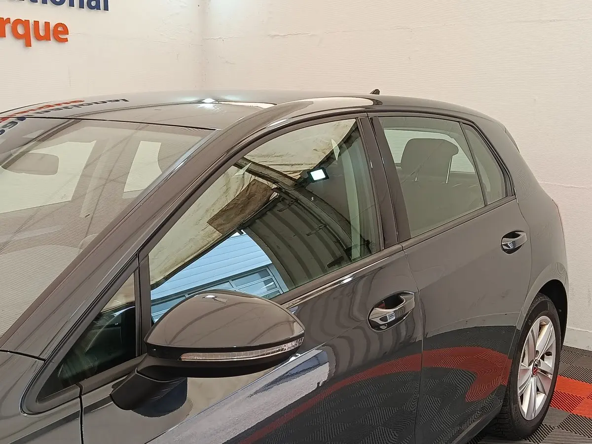 Vue latérale côté gauche d'une Volkswagen Golf gris foncé avec rétroviseur et poignées de porte visibles dans un showroom.