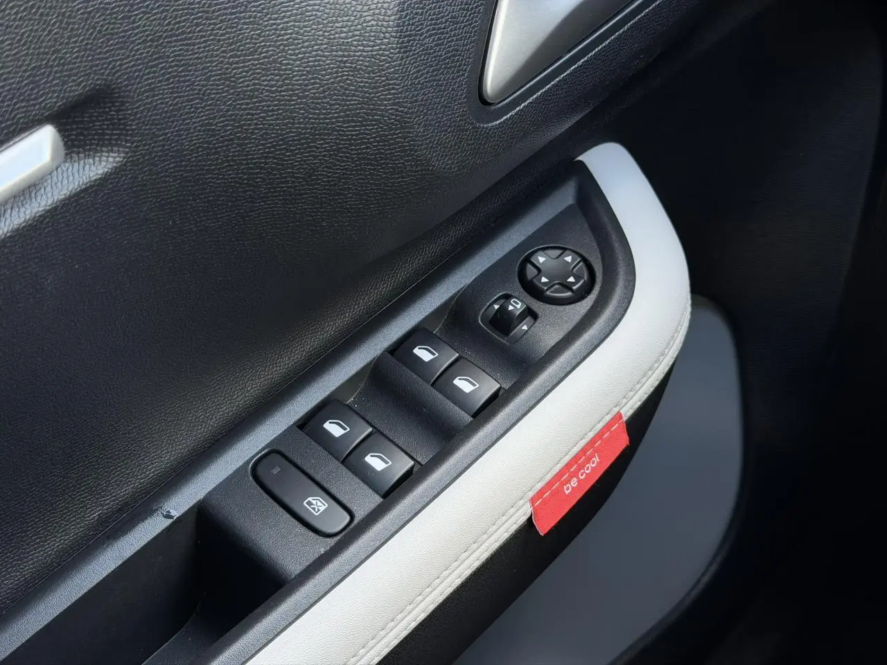 Gros plan sur la commande des vitres électriques côté conducteur avec surpiqûres blanches et étiquette rouge "be cool" sur la portière.