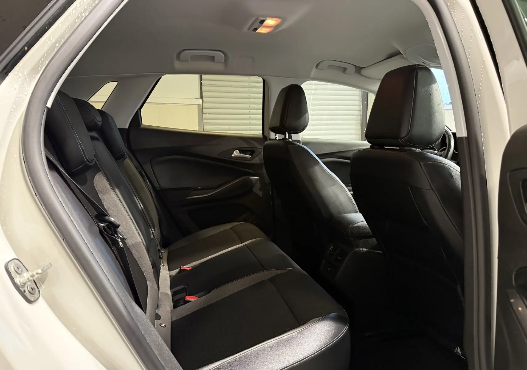 Vue intérieure arrière gauche de l’Opel Grandland X blanc, montrant les sièges noirs en cuir et tissu, avec éclairage plafond allumé.