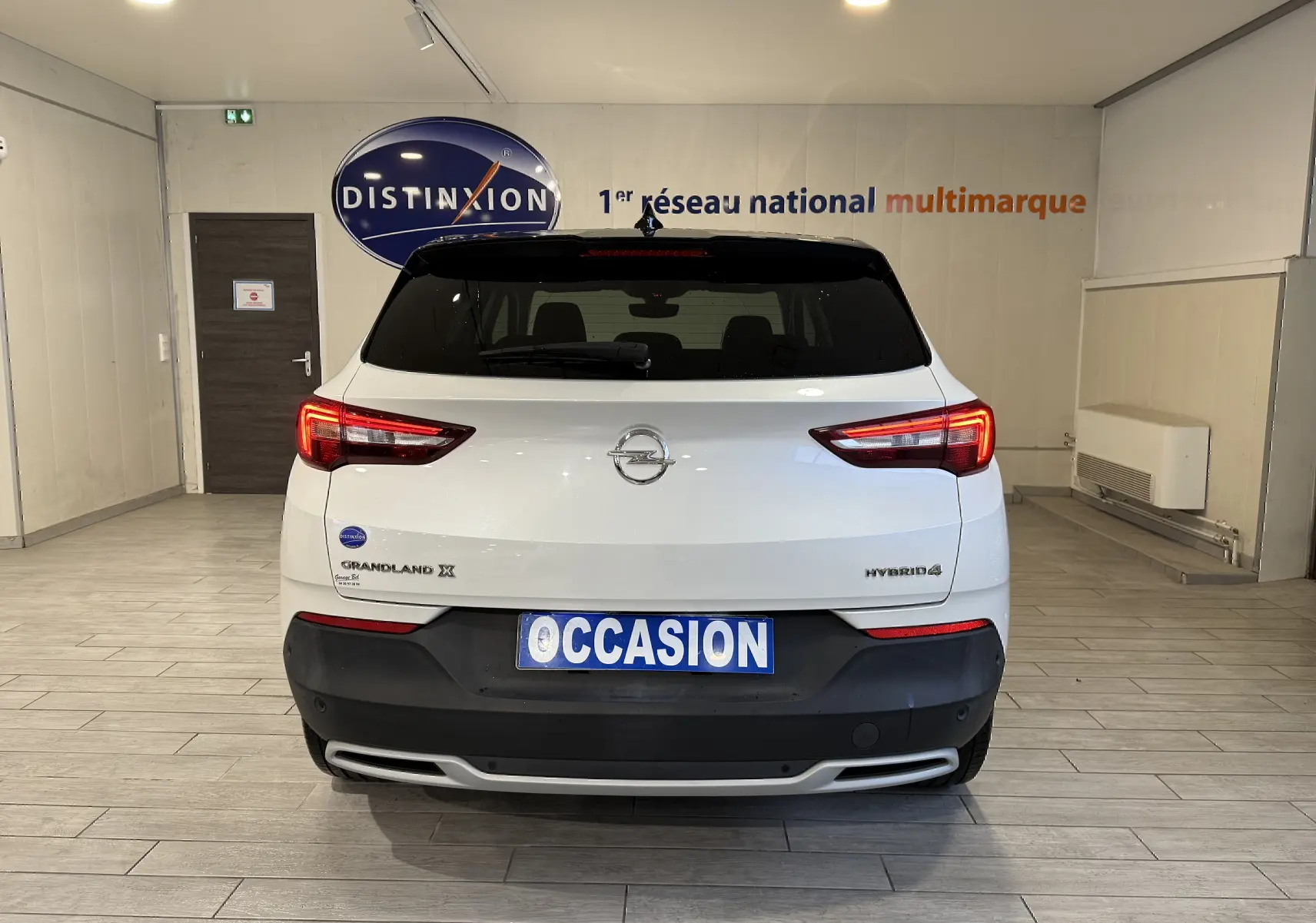 Vue arrière d'un Opel Grandland X blanc hybride rechargeable, avec feux allumés et plaque "OCCASION" en intérieur showroom.