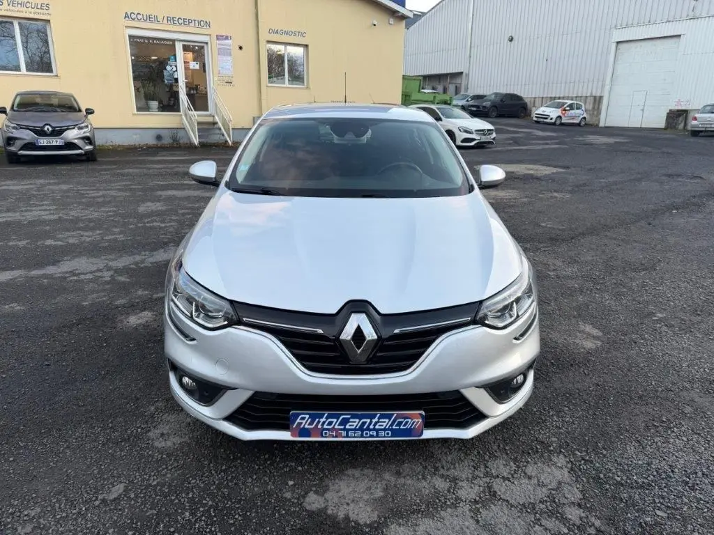 Vue avant d'une Renault Mégane Business gris alu avec calandre noire et phares LED, stationnée devant un bâtiment jaune.