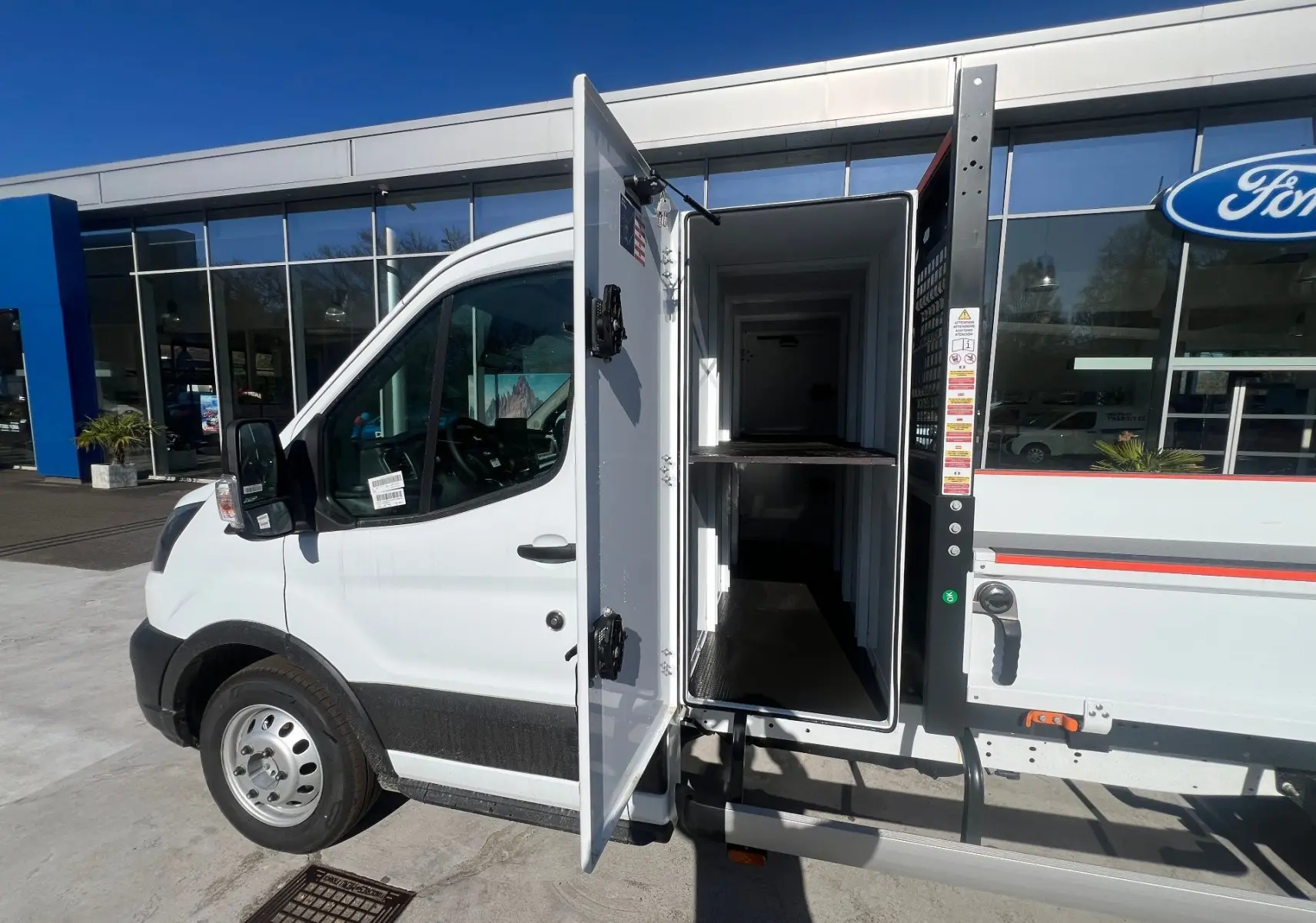 Vue de profil côté gauche d’un Ford Transit blanc 2026 avec porte latérale ouverte montrant la benne et le coffre intégré.
