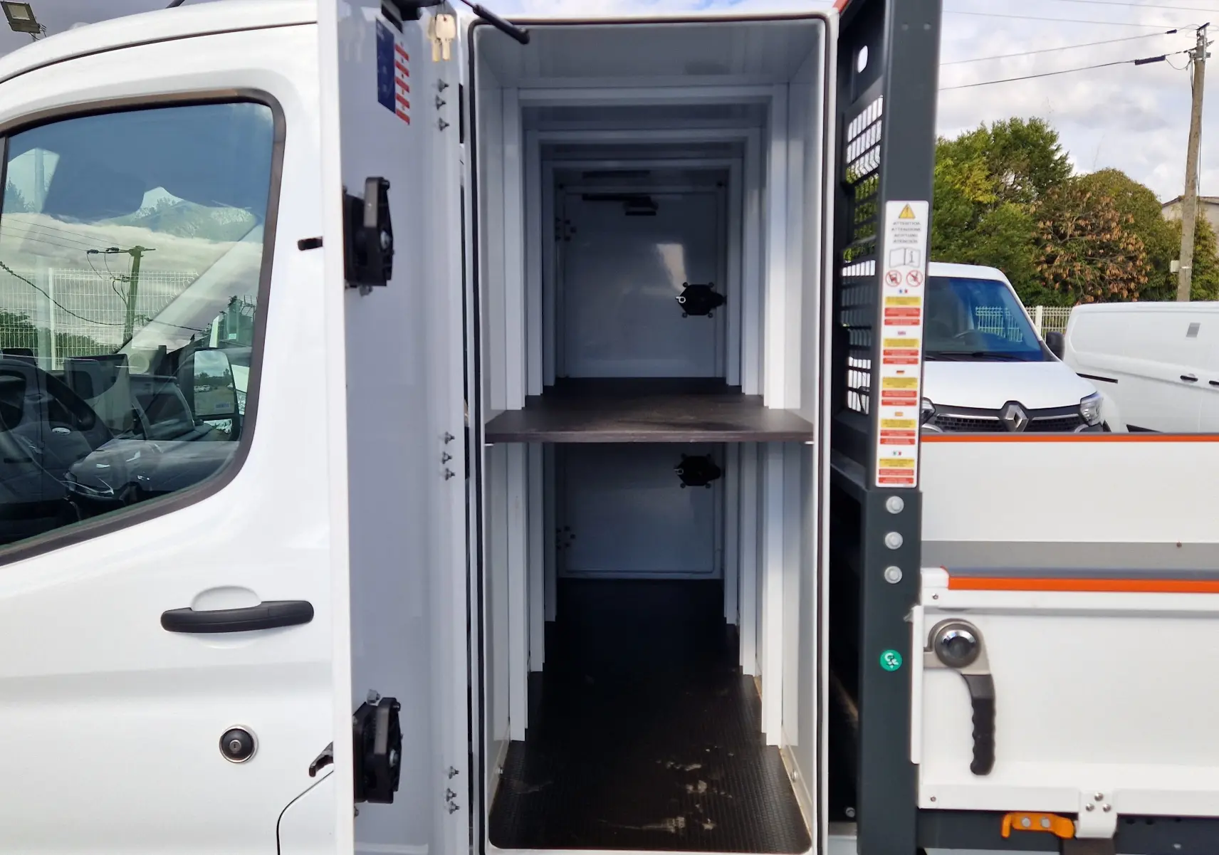 Vue latérale du Ford Transit Chassis Cabine blanc 2026 avec porte ouverte montrant le coffre de rangement intégré à la benne acier.