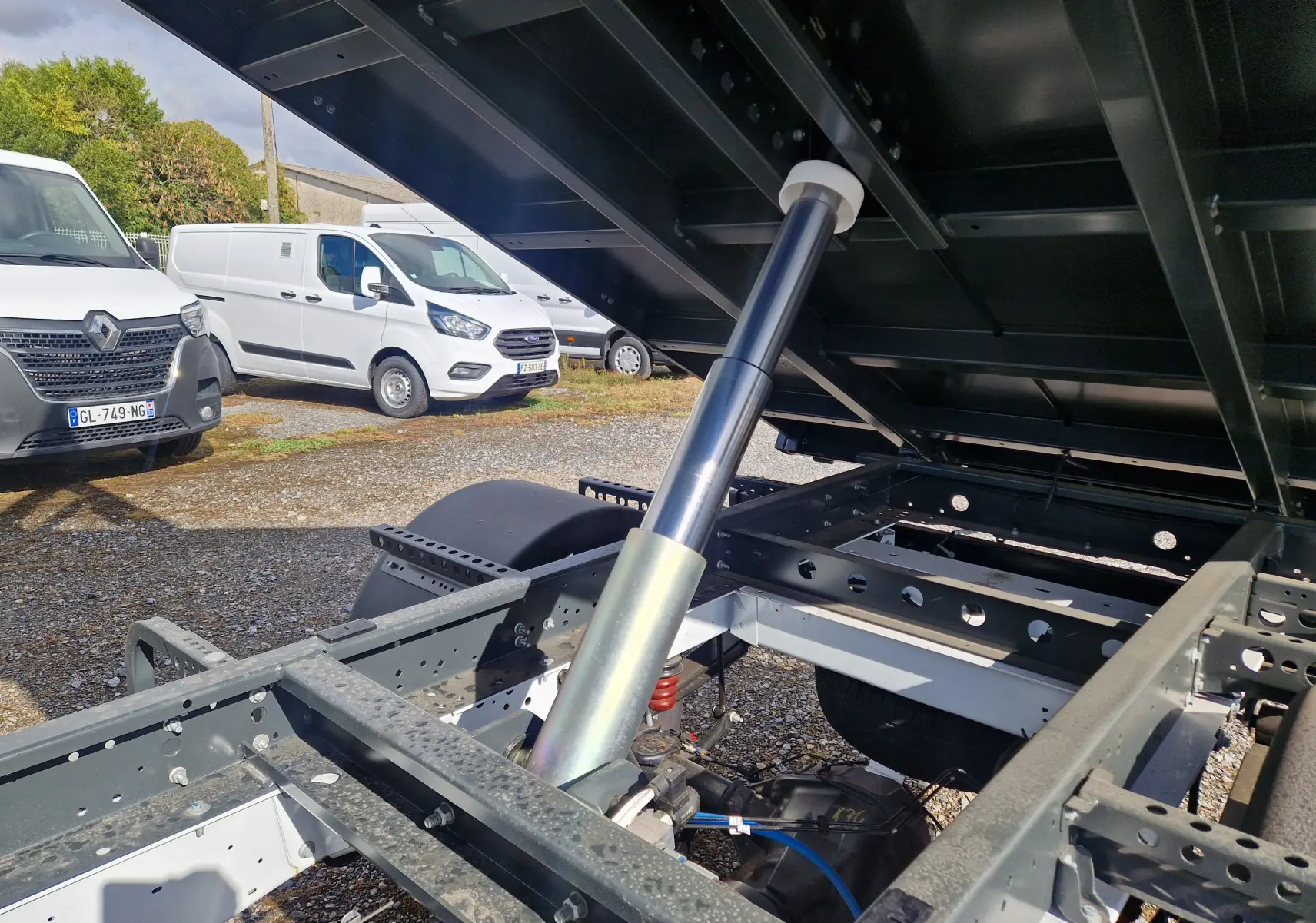 Vue rapprochée sous la benne relevée d’un Ford Transit blanc, mettant en valeur le vérin hydraulique et le châssis acier.