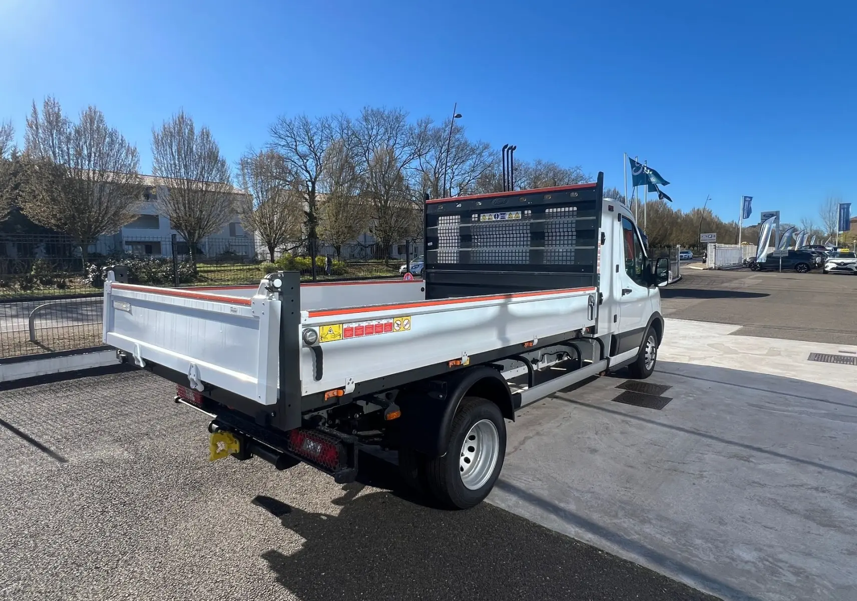 Vue 3/4 arrière droite d’un Ford Transit Châssis Cabine blanc avec benne et coffre, roues jumelées, sur parking extérieur.