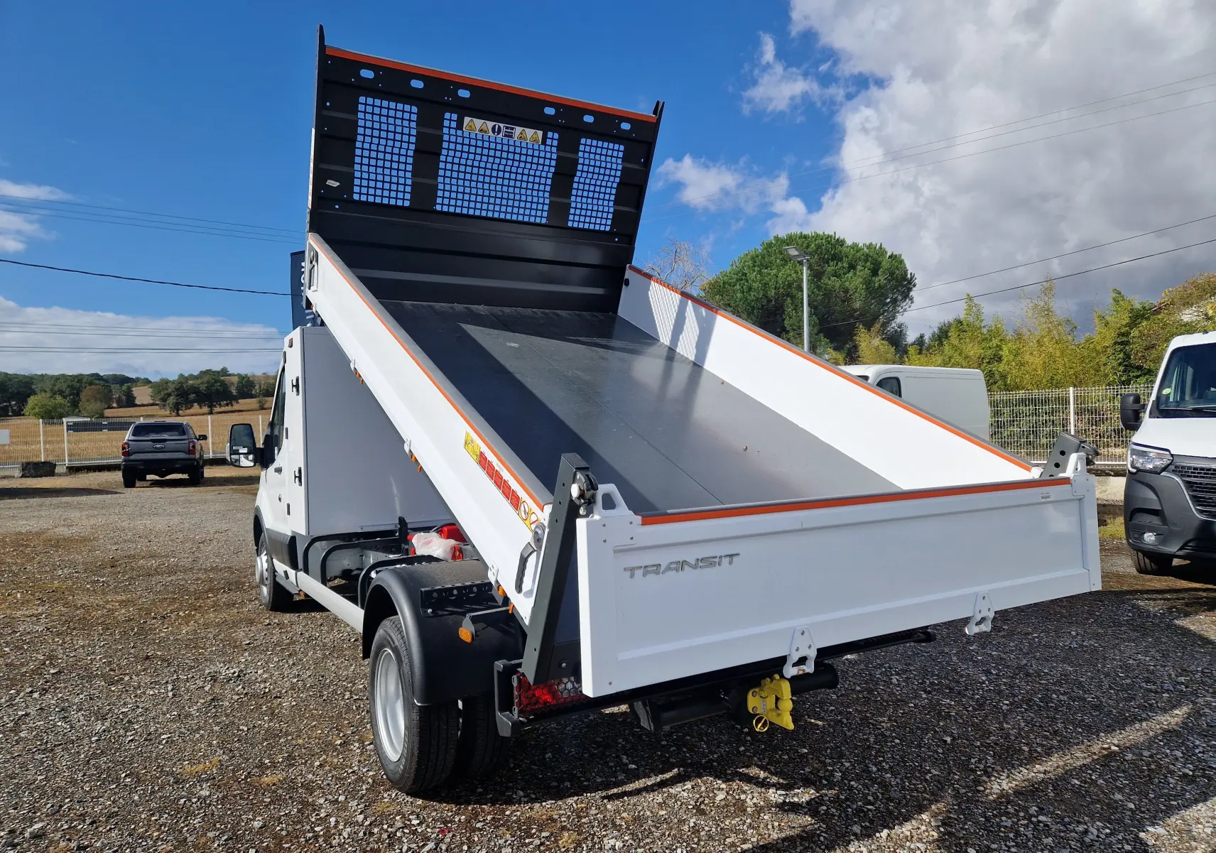 Vue 3/4 arrière droite du Ford Transit blanc avec benne relevée et coffre latéral visible sur terrain extérieur.
